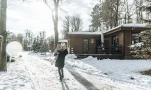 Kobieta z dzieckiem na rękach idzie zimową drogą do drewnianego domku.